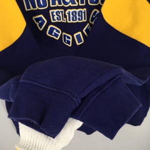 May include: Navy blue and yellow sweatshirt with the text "NORTHGATE" and "EST. 1891". The sweatshirt has yellow panels on the shoulders and a ribbed cuff. The letters "AGGIE" are also visible.