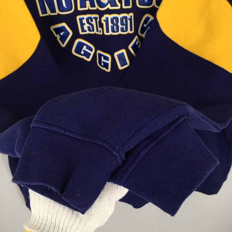 May include: Navy blue and yellow sweatshirt with the text "NORTHGATE" and "EST. 1891". The sweatshirt has yellow panels on the shoulders and a ribbed cuff. The letters "AGGIE" are also visible.