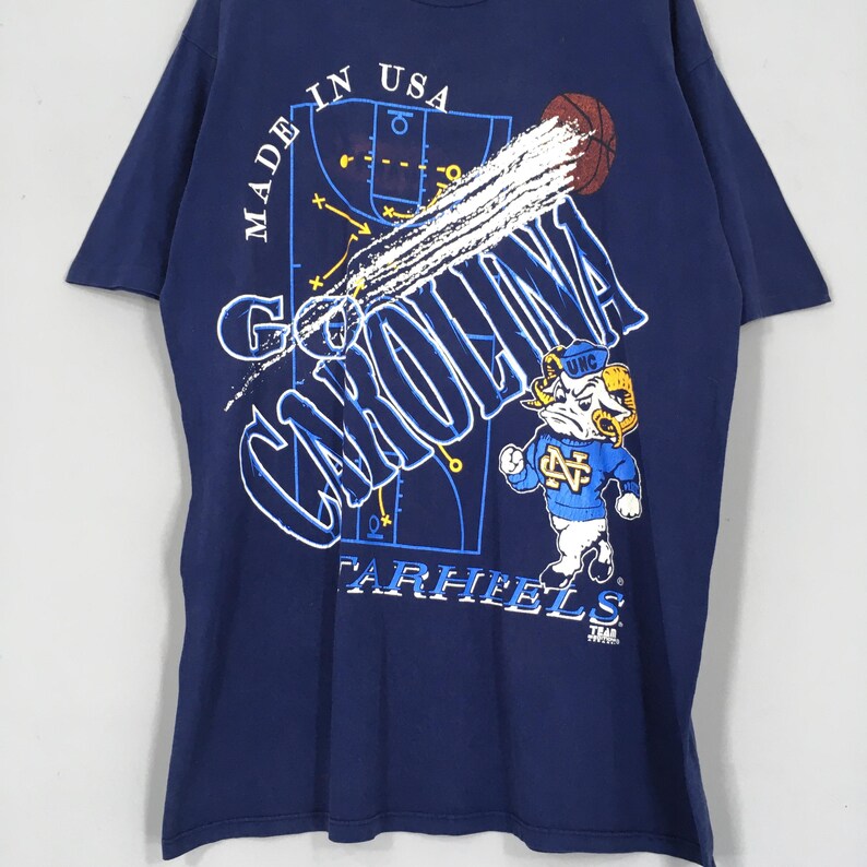 May include: Navy blue t-shirt with a basketball graphic and the words "Go Carolina" in white. The shirt features a cartoon ram mascot wearing a blue sweater and a basketball court design. Made in USA.