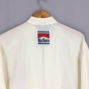 May include: A cream-colored long-sleeve shirt featuring a rectangular patch on the upper back. The patch displays the text HONDA in black, a red chevron shape, the word Marlboro in black, and the word McLaren in black on a white background.