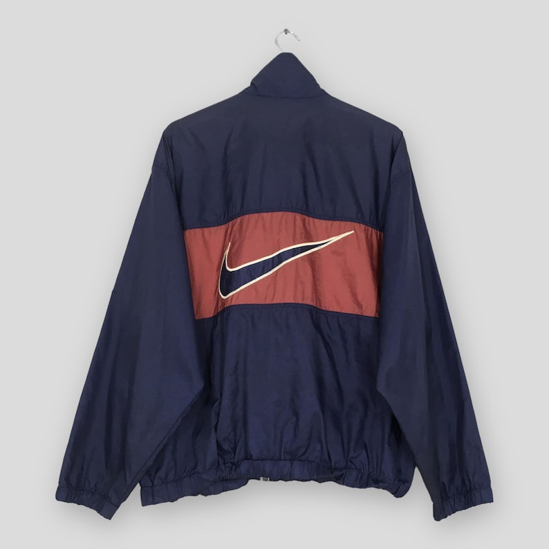 Pu&ograve; includere: Giacca a vento blu navy con una striscia rossa sulla schiena con il logo Nike bianco con un contorno blu navy. La giacca ha un collo alto e una cerniera a tutta lunghezza.