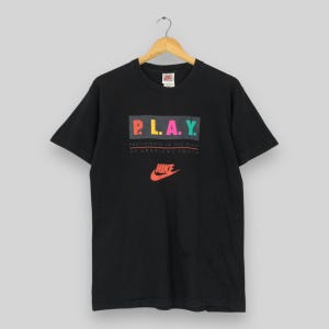 Puede incluir: Camiseta negra con la palabra "P.L.A.Y." en letras coloridas, encima del texto "Participate in the lives of American youth" y el logo de Nike en naranja.