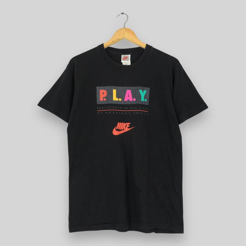 Puede incluir: Camiseta negra con la palabra "P.L.A.Y." en letras coloridas, encima del texto "Participate in the lives of American youth" y el logo de Nike en naranja.