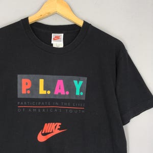 Puede incluir: Camiseta negra con la palabra "PLAY" en letras coloridas. Debajo, el texto dice "PARTICIPATE IN THE LIVES OF AMERICA'S YOUTH". El logotipo de Nike est&aacute; en rojo debajo.