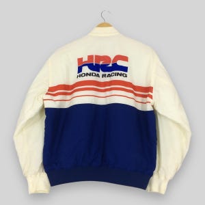 May include: A white, red, and blue long-sleeve jacket featuring the HRC Honda Racing logo on the back. The design includes a large blue panel at the bottom, horizontal red stripes across the middle, and a white upper section with long sleeves.