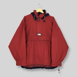 May include: A red pullover jacket with a dark blue hood lining and collar. It features a front pouch pocket, a partial button placket, and a rectangular patch on the chest with the text CHAPS RALPH LAUREN. The cuffs have adjustable straps.