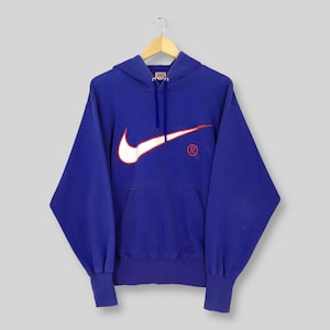 May include: A blue hoodie with a white Nike swoosh logo and a red registered trademark symbol. The hoodie has a drawstring hood and a kangaroo pocket. The sweatshirt is hanging on a wooden hanger.