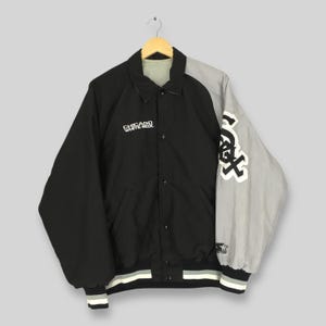 May include: A black and grey Chicago White Sox jacket with the team logo on the sleeve. The jacket features a button-up front, a collar, and the words "Chicago White Sox" printed on the chest. The jacket has a black body and a grey sleeve.