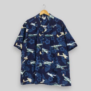 May include: A short-sleeved navy blue button-down shirt featuring a repeating pattern of vintage propeller aeroplanes and dark blue hibiscus flowers. The shirt has a classic collar, a front button placket, and is displayed on a wooden hanger.