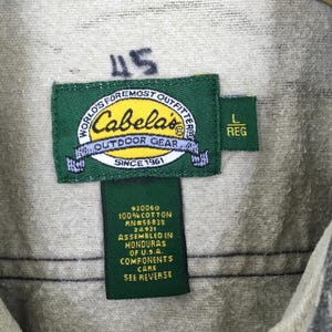 Puede incluir: Primer plano de una etiqueta de prenda con el logotipo de Cabela's, un rect&aacute;ngulo verde con texto blanco. La etiqueta tambi&eacute;n incluye el texto "WORLD'S FOREMOST OUTFITTERS" y "OUTDOOR GEAR". Etiquetas adicionales indican la talla "L REG" y la composici&oacute;n de la tela.