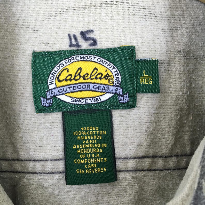 Puede incluir: Primer plano de una etiqueta de prenda con el logotipo de Cabela's, un rect&aacute;ngulo verde con texto blanco. La etiqueta tambi&eacute;n incluye el texto "WORLD'S FOREMOST OUTFITTERS" y "OUTDOOR GEAR". Etiquetas adicionales indican la talla "L REG" y la composici&oacute;n de la tela.