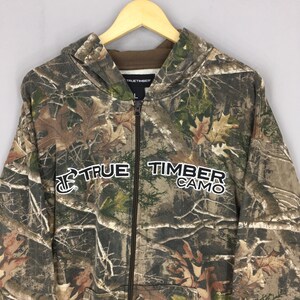 May include: A camouflage zip-up hoodie with the text "TRUE TIMBER CAMO" in white. The hoodie features a brown interior lining and a hood. The pattern includes shades of brown, green, and tan, resembling a forest.