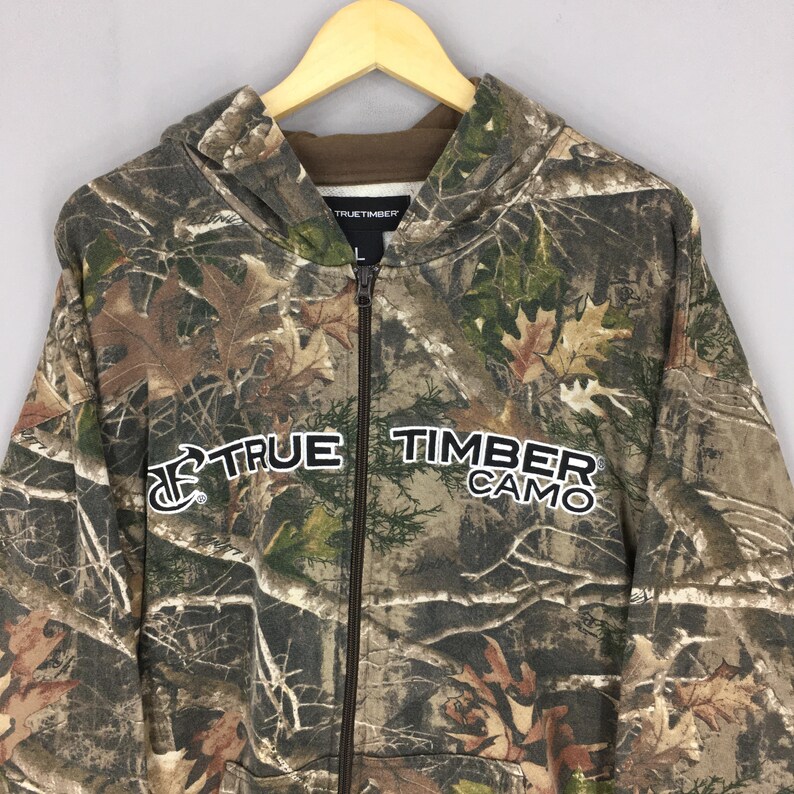May include: A camouflage zip-up hoodie with the text "TRUE TIMBER CAMO" in white. The hoodie features a brown interior lining and a hood. The pattern includes shades of brown, green, and tan, resembling a forest.