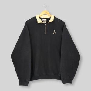 May include: Black pullover sweatshirt with a cream-colored collar and a quarter-zip closure. The sweatshirt features a small embroidered Mickey Mouse design on the chest. The garment is hanging from a wooden hanger.