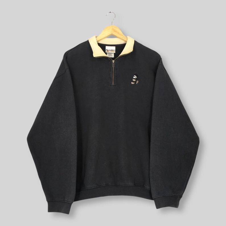May include: Black pullover sweatshirt with a cream-colored collar and a quarter-zip closure. The sweatshirt features a small embroidered Mickey Mouse design on the chest. The garment is hanging from a wooden hanger.