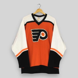 May include: An orange and black hockey jersey featuring the Philadelphia Flyers logo. The jersey has white mesh sleeves with black and orange detailing. The V-neck collar is black.