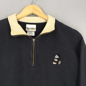 May include: A black sweatshirt with a cream-colored collar and a quarter-zip closure. The sweatshirt features an embroidered Mickey Mouse design on the front and a "Walt Disney World" tag. The garment is hanging on a wooden hanger.