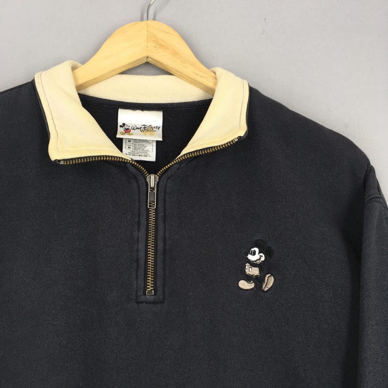 May include: A black sweatshirt with a cream-colored collar and a quarter-zip closure. The sweatshirt features an embroidered Mickey Mouse design on the front and a "Walt Disney World" tag. The garment is hanging on a wooden hanger.