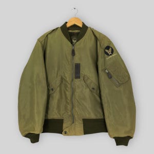 May include: An olive green bomber jacket featuring a full-length front zipper, ribbed cuffs and hem, and two front pockets with snap closures. A circular U.S. Air Force patch is on the left sleeve above a zippered utility pocket with a pull tab.