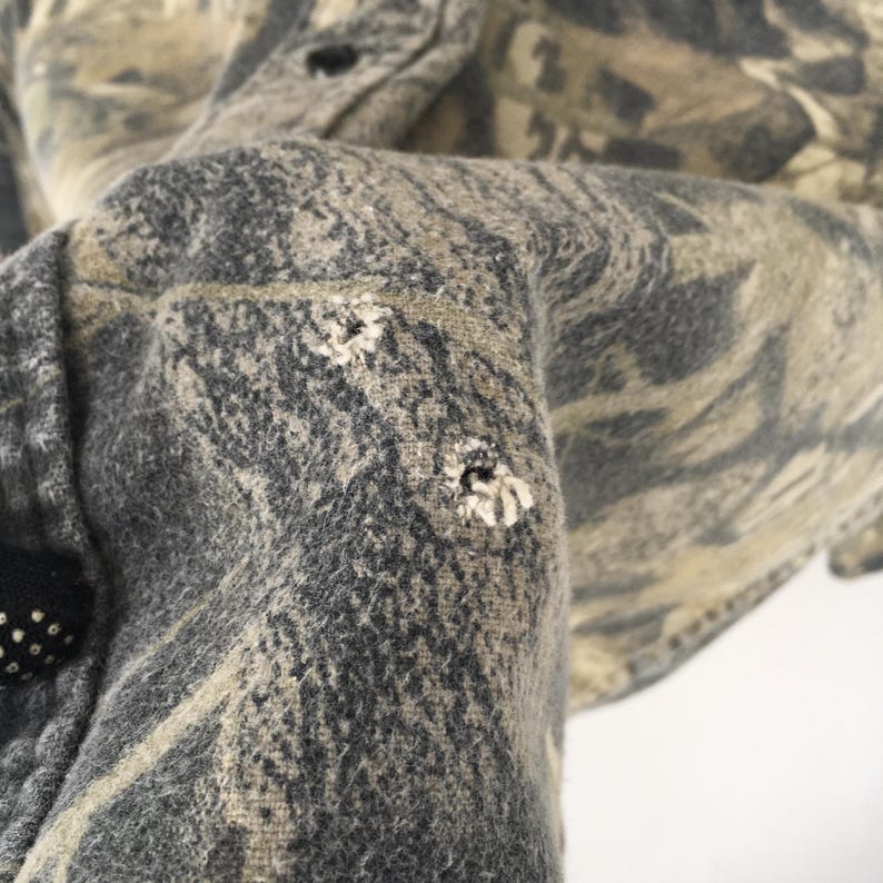 May include: Close-up of a camouflage-patterned shirt sleeve. The fabric features a blend of gray, brown, and tan hues. The sleeve has several small holes, indicating wear and tear. The shirt is buttoned at the cuff.