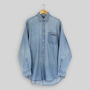 May include: Light blue denim button-down shirt with long sleeves. Features a collared neckline, a chest pocket with a small logo, and buttoned cuffs. The shirt is hanging on a wooden hanger.