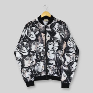 May include: A zip-up bomber jacket featuring an all-over black and white graphic print of multiple stylized human faces. The jacket has a black ribbed collar, black ribbed cuffs, and a black ribbed hem, with a central black zipper closure.