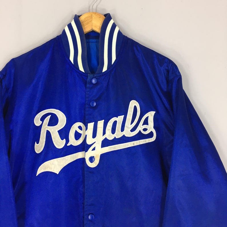 Pu&ograve; includere: Giacca in raso blu reale con la scritta "Royals" in bianco sul davanti. La giacca ha un colletto a righe bianche e blu e un'abbottonatura. La giacca &egrave; appesa a una gruccia di legno.