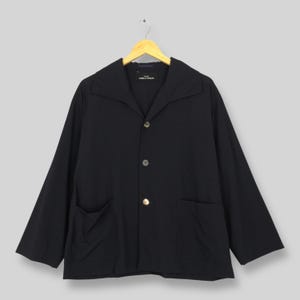 Pode incluir: Um casaco preto com gola larga e três botões. O casaco tem dois bolsos frontais e mangas compridas. O casaco está pendurado num cabide de madeira sobre um fundo cinza claro. A etiqueta diz "COMME des GARÇONS".