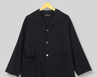 May include: A black jacket with a wide collar and three buttons. The jacket has two front pockets and long sleeves. The jacket is hanging on a wooden hanger against a light gray background. The label reads "COMME des GARÇONS".