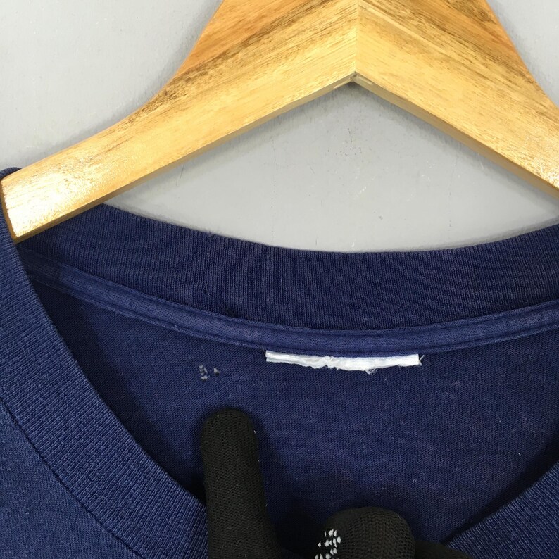 May include: Close-up of a navy blue t-shirt with a V-neck collar. The shirt is hanging on a wooden hanger. A white tag is visible near the collar.