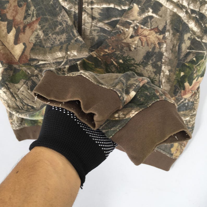May include: Camouflage zip-up hoodie with brown cuffs. The jacket features a woodland camouflage pattern in shades of brown, green, and tan. The cuffs are a solid brown colour.