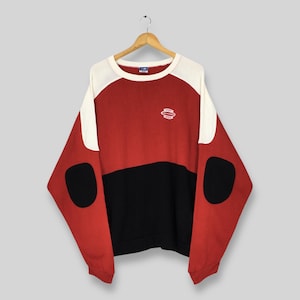 Puede incluir: Una sudadera con bloques de color en rojo, negro y blanco, con un pequeño logotipo en el pecho. La parte superior de manga larga tiene cuello redondo y un ajuste relajado. La sudadera está colgada en una percha de madera.