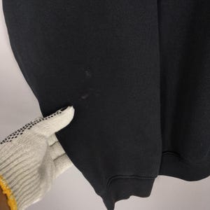 May include: Close-up of a black sweatshirt. The fabric appears to be a thick knit. There are a few small, dark spots on the sleeve. The cuff is ribbed. A gloved hand is touching the sleeve.