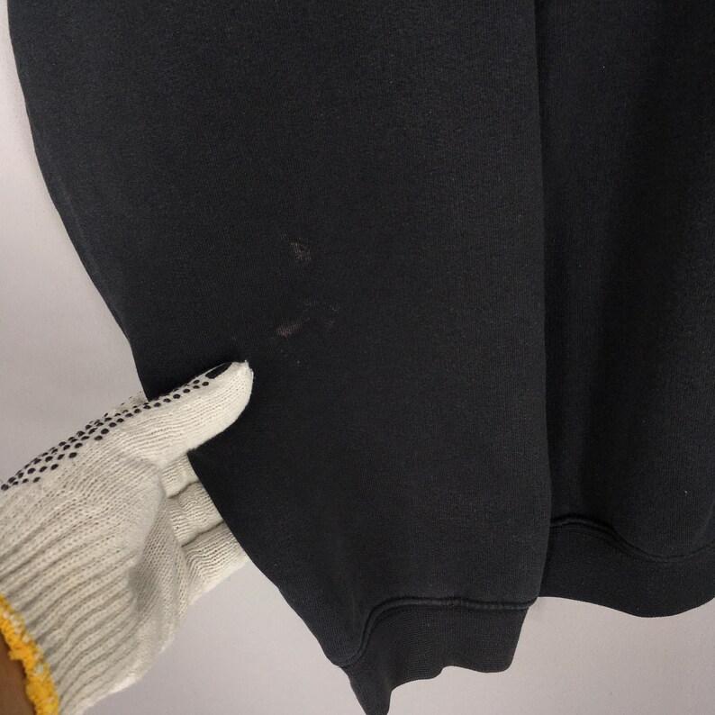 May include: Close-up of a black sweatshirt. The fabric appears to be a thick knit. There are a few small, dark spots on the sleeve. The cuff is ribbed. A gloved hand is touching the sleeve.