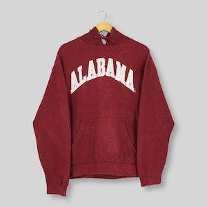 May include: A maroon hoodie with the word "ALABAMA" arched across the chest in white block letters. The hoodie has a front pocket and a drawstring hood. The garment is hanging on a wooden hanger.