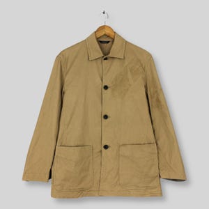 Op de afbeelding: Een beige jas met knoopsluiting, voorzien van een klassieke kraag, lange mouwen en twee grote opgestikte zakken aan de voorzijde. Het kledingstuk wordt gesloten met drie donkere knopen in het midden en heeft een klein label in de nek.