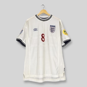 Może przedstawiać: Biała koszulka piłkarska reprezentacji Anglii z granatowymi wykończeniami i numerem 8 w kolorze bordowym. Zawiera herb Anglii i logo Umbro. Wisząca na drewnianym wieszaku.