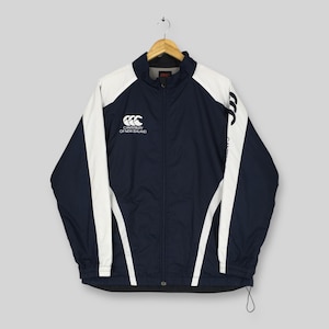 May include: A navy blue and white zip-up windbreaker jacket featuring the Canterbury of New Zealand logo printed in white on the right chest and a vertical Canterbury logo along the left sleeve, with a high collar and long sleeves.