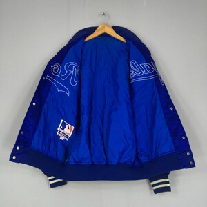 Pu&ograve; includere: Giacca da baseball blu e bianca con chiusura a scatto. La giacca presenta scritte bianche e un logo della Major League Baseball. I polsini e il colletto sono blu navy con strisce bianche.