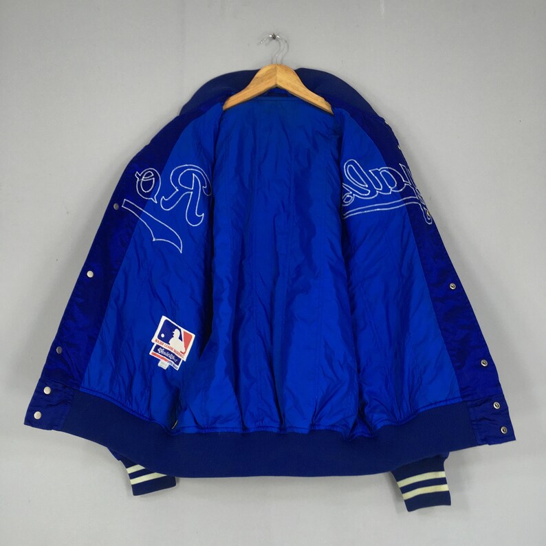 Pu&ograve; includere: Giacca da baseball blu e bianca con chiusura a scatto. La giacca presenta scritte bianche e un logo della Major League Baseball. I polsini e il colletto sono blu navy con strisce bianche.