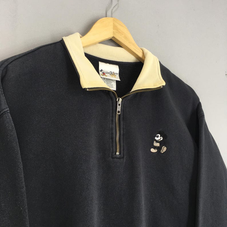 May include: A black zip-up sweatshirt with a cream-colored collar and a small embroidered Mickey Mouse design. The sweatshirt has a half-zip closure and a tag with the Disney logo.