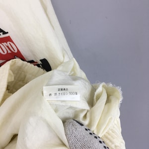 May include: An off-white nylon jacket featuring a partial red and black graphic print. A white interior care label is visible, displaying the Japanese text 品質表示 and 表地 ナイロン 100%, indicating the outer material is 100% nylon.