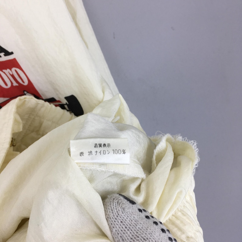 May include: An off-white nylon jacket featuring a partial red and black graphic print. A white interior care label is visible, displaying the Japanese text 品質表示 and 表地 ナイロン 100%, indicating the outer material is 100% nylon.