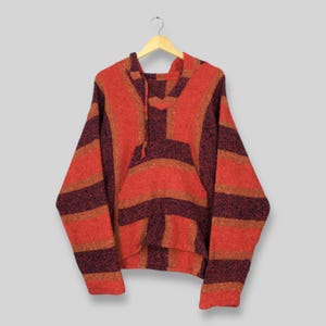 May include: A red and burgundy striped Baja hoodie with a hood and front pocket. The woven fabric has a textured appearance. The hoodie is hanging on a wooden hanger against a neutral background. The garment is a pullover style.