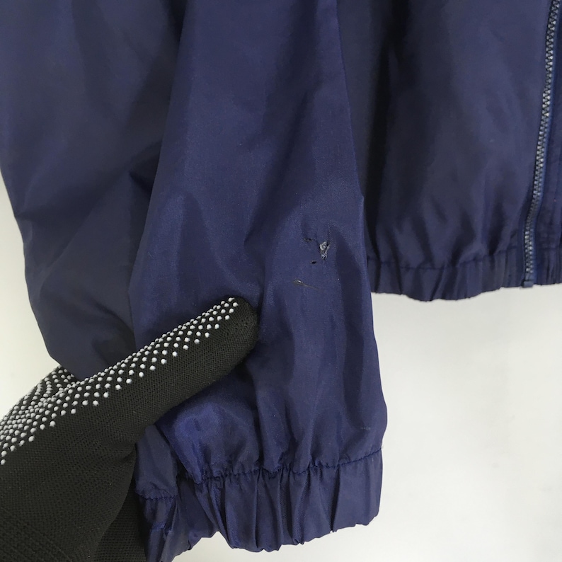 Pu&ograve; includere: Primo piano di una giacca blu navy con cerniera intera. La giacca ha orlo e polsini arricciati. &Egrave; visibile un piccolo strappo sulla manica sinistra. La giacca &egrave; realizzata in un materiale leggero.