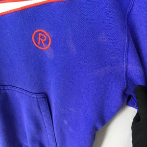 May include: Close-up of a blue sweatshirt with a white and orange stripe across the top. The sweatshirt has a kangaroo pocket and a circular orange logo with the letter "R".