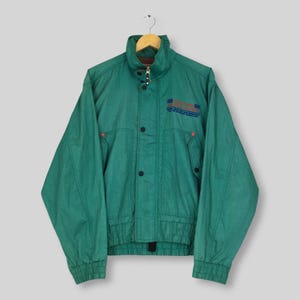May include: A forest green windbreaker jacket featuring a high collar, long sleeves with elastic cuffs, and a front zipper closure with a snap-button placket. The chest displays blue and red embroidered text and a small logo graphic.