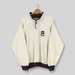 May include: Cream-colored pullover sweatshirt with a brown collar, cuffs, and hem. The sweatshirt features a quarter-zip closure and a small embroidered emblem with a maple leaf and the text "Niagara Falls Canada".