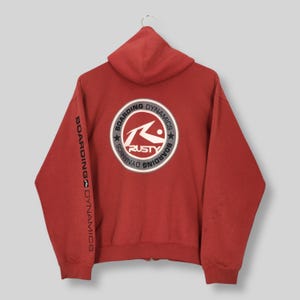 May include: Red zip-up hoodie with a hood and long sleeves. The back features a circular logo with the text "RUSTY" and "BOARDING DYNAMICS". The left sleeve has the text "BOARDING DYNAMICS" printed vertically.
