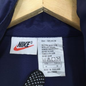Pu&ograve; includere: Primo piano di un'etichetta di abbigliamento blu navy con il logo Nike in rosso e bianco. L'etichetta include le istruzioni di lavaggio, la taglia M e il testo "MADE IN THAILAND RN #56323". L'indumento &egrave; realizzato in 100% nylon.